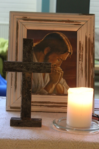 Pöydällä risti ja kynttilä, joiden takana taulu, jossa Jeesu rukoilee.